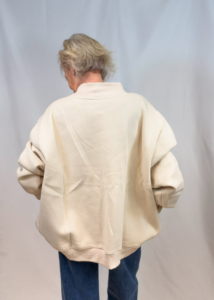 Woman shown from back wearing cream color mock neck sweatshirt and jeans on plain background