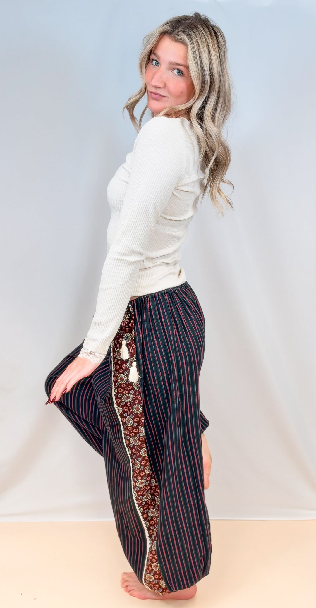 Woman shown from side view wearing cream color long sleeve top with black, green and rose color pin stripe balloon pants on plain background