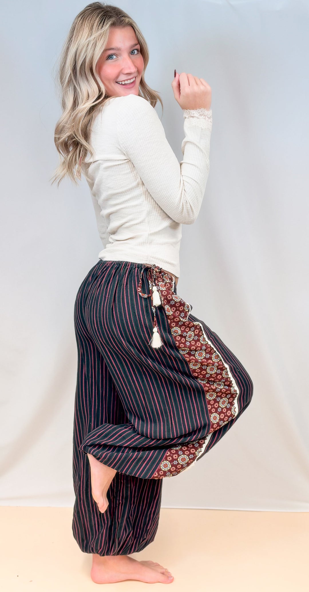 Woman shown from side view wearing cream color long sleeve top with black, green and rose color stripe balloon pants on plain background