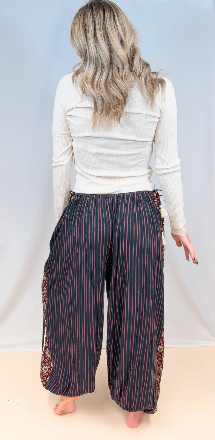 Woman shown from back view wearing cream color long sleeve top with black, green and rose color stripe balloon pants on plain background