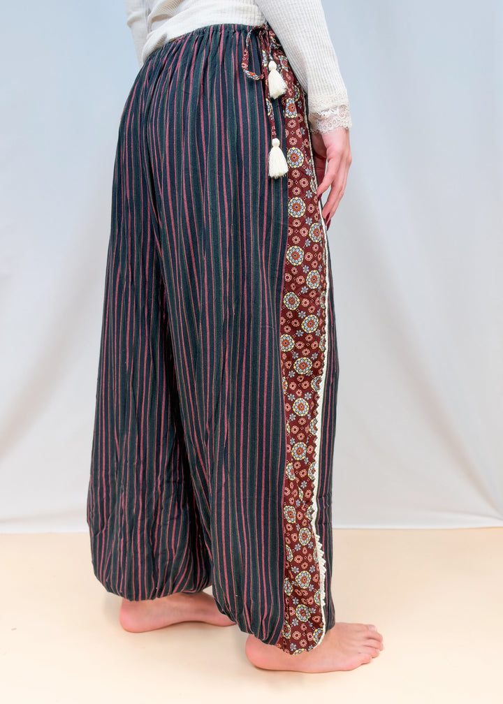 Woman shown from side view wearing black, green and rose color pin stripe balloon pants on plain background
