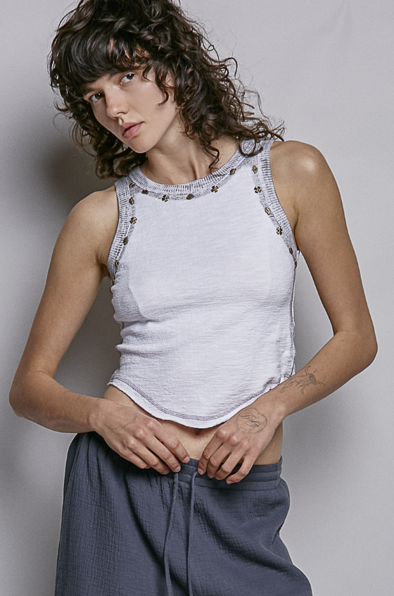 A model wearing a light gray ribbed knit tank top with silver metallic studs along the neckline, paired with dark gray pants, against a neutral gray background.
