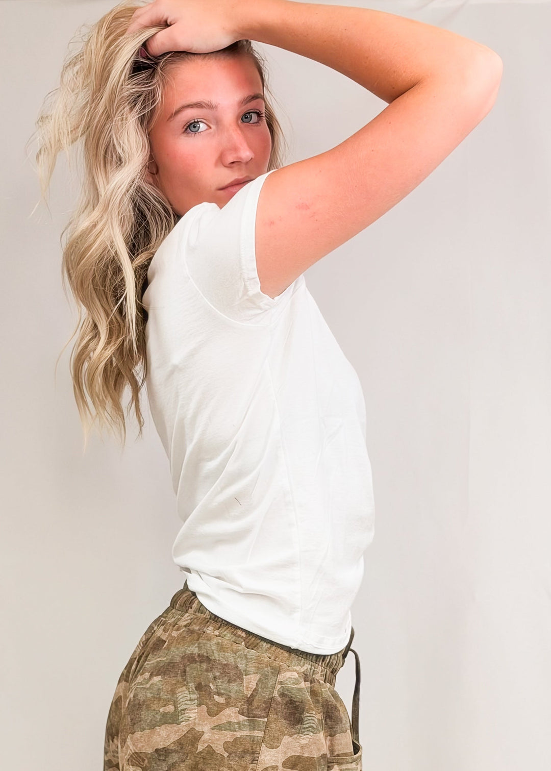 Woman wearing white baby tee with camo pants on plain background