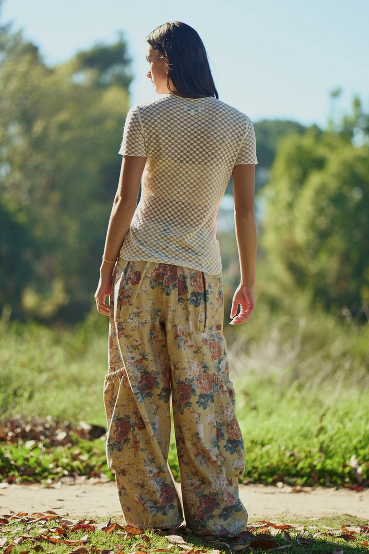Woman wearing a patterned top and floral pants standing in a natural setting with trees and grass.
