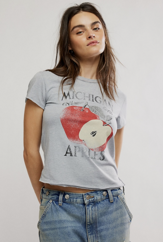 A woman wearing a light gray graphic t-shirt with a red apple print and the words &