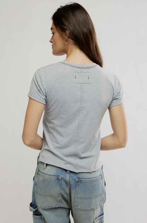 A woman viewed from the back wearing a light gray short-sleeve t-shirt with a small back pocket detail and light blue jeans, set against a plain light gray studio background.