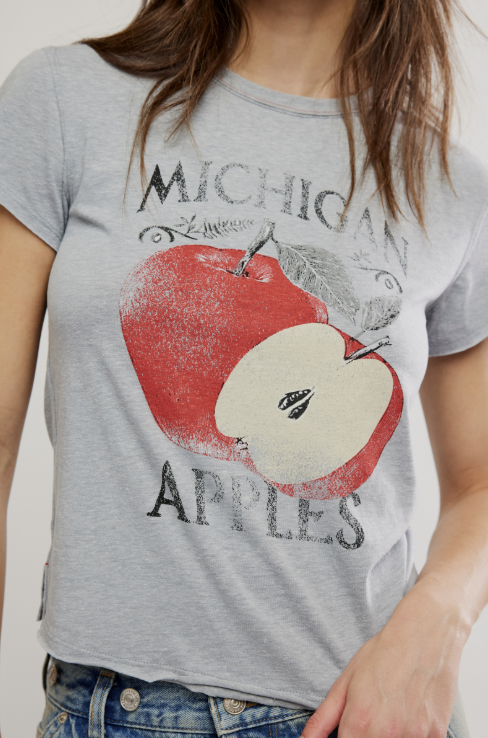 A woman wearing a light gray heather graphic t-shirt featuring a distressed red and white apple design with the text &