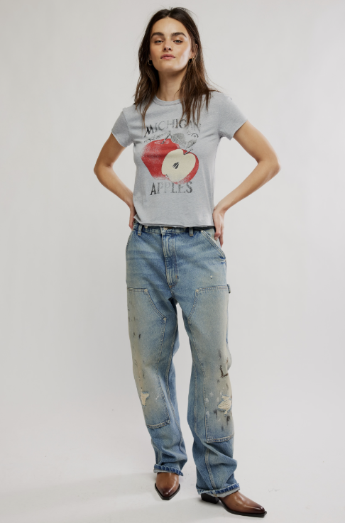 A woman modeling a light gray graphic t-shirt with a red apple print and distressed light blue jeans, standing against a plain light gray studio background.