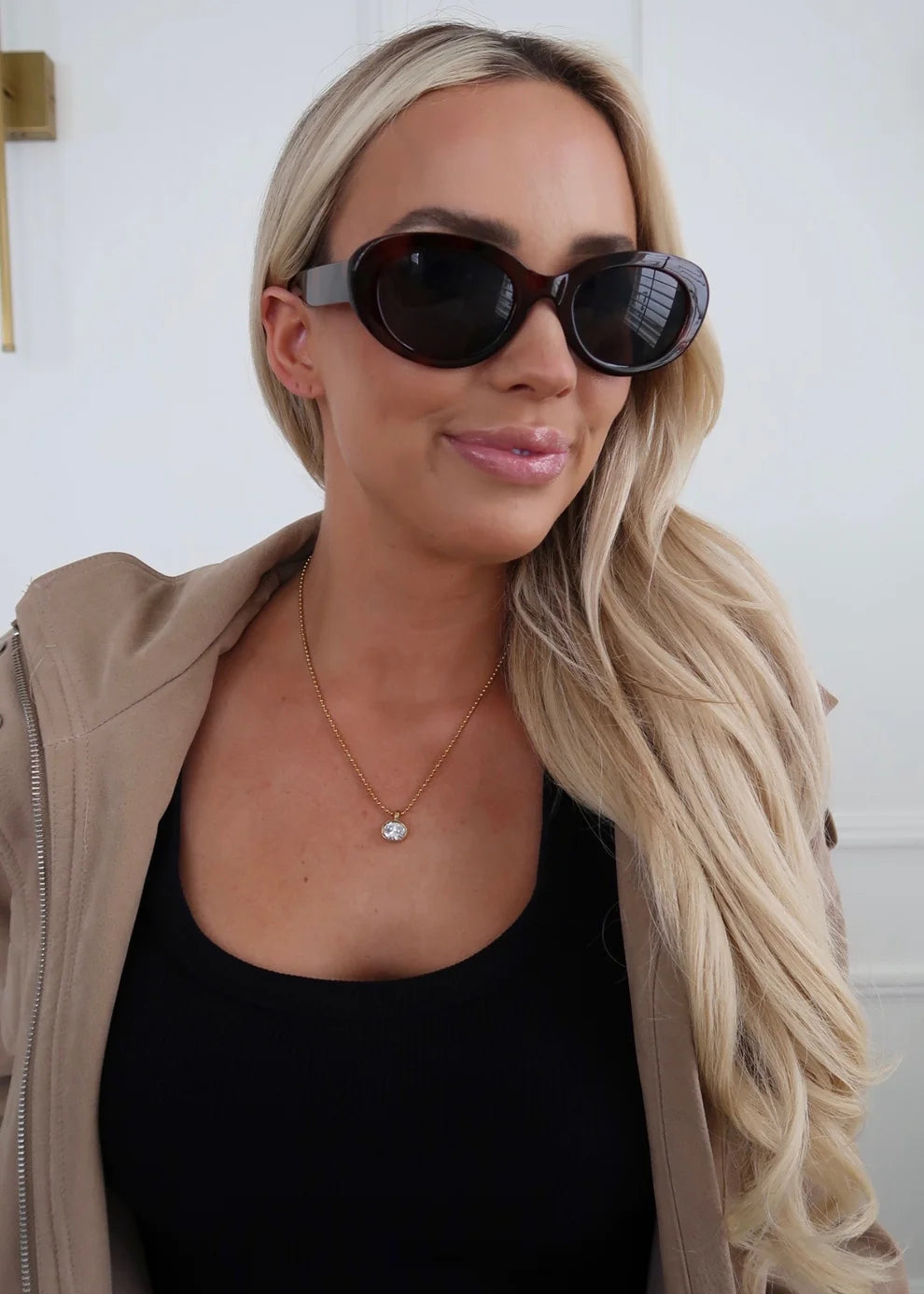 A woman with long blonde hair wearing dark brown cat-eye sunglasses, a black top, and a beige zip-up jacket. She is wearing a delicate gold necklace with a small heart pendant.
