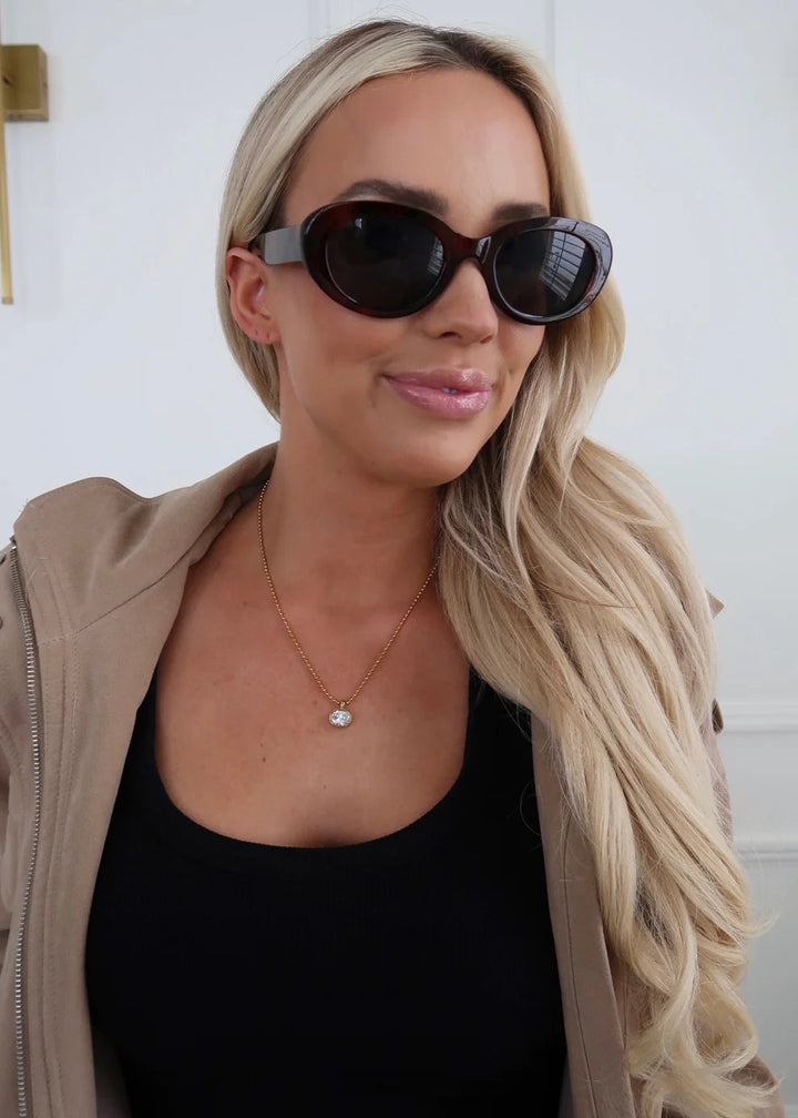 A woman with long blonde hair wearing dark brown cat-eye sunglasses, a black top, and a beige zip-up jacket. She is wearing a delicate gold necklace with a small heart pendant.