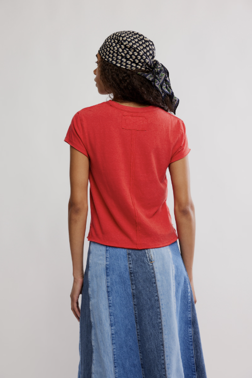 Person wearing a red t-shirt and blue denim skirt with a patterned headscarf on a plain background