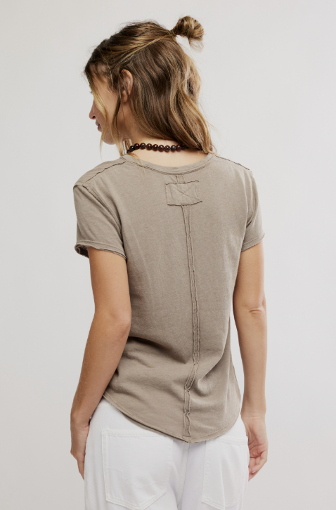 A woman shown from the back wearing a taupe short-sleeve t-shirt with a vertical seam detail and white pants, against a light gray studio background.