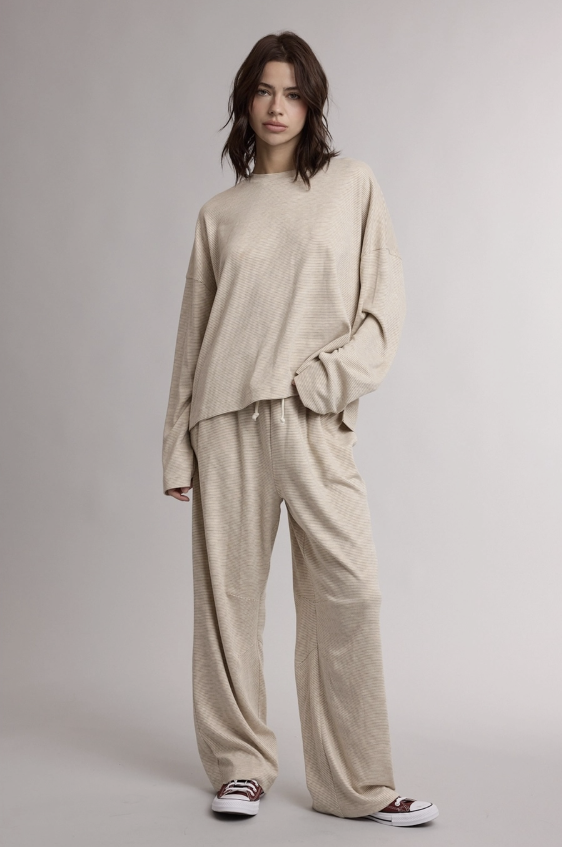 A model wearing a matching beige lounge set consisting of a long-sleeve cropped sweatshirt and wide-leg sweatpants, paired with black and white sneakers, against a neutral gray studio background.