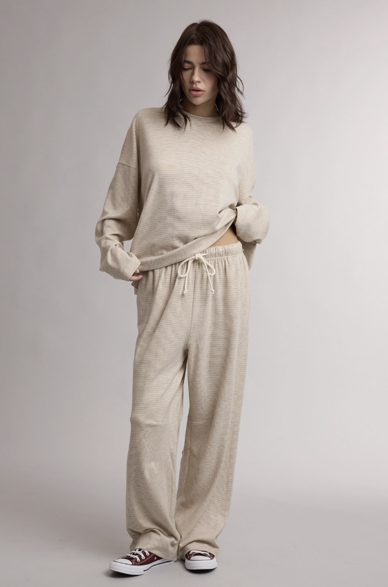 A woman modeling a matching beige knit lounge set consisting of a long-sleeve crewneck sweater and wide-leg drawstring pants, paired with white sneakers, against a neutral gray studio background.