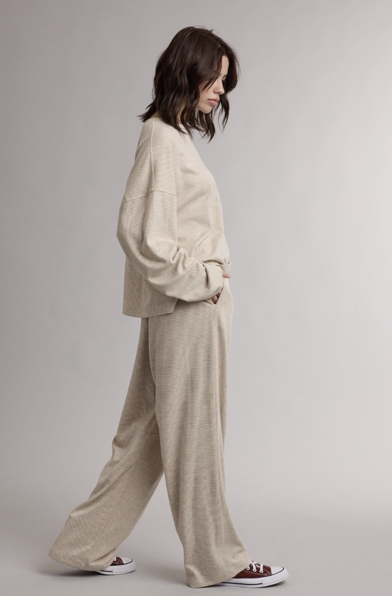 A woman wearing a matching beige lounge set consisting of a cropped long-sleeve top and wide-leg pants, shown in profile against a neutral gray studio background.