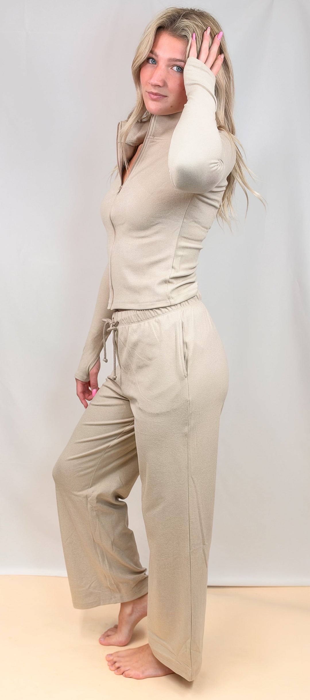 A woman modeling a beige two-piece loungewear set consisting of a long-sleeve mock-neck top and matching wide-leg pants, standing against a light gray studio backdrop.