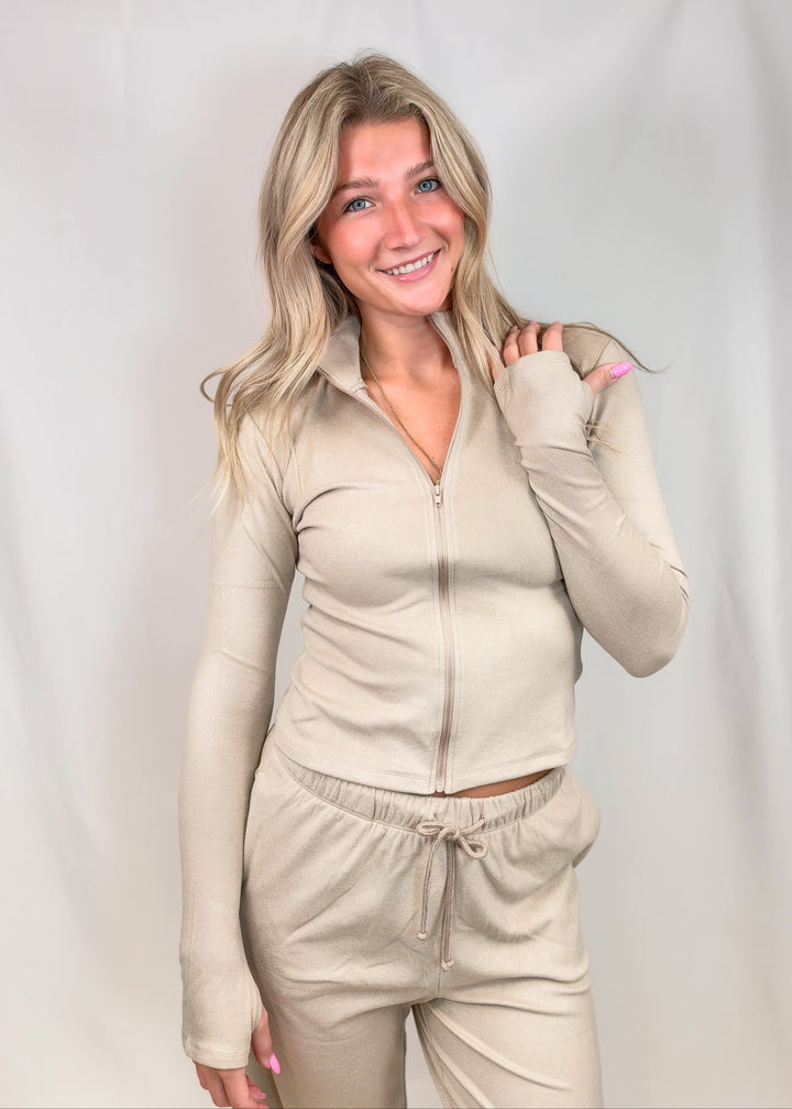 A woman modeling a matching beige zip-up long-sleeve top and drawstring sweatpants against a light gray studio backdrop.