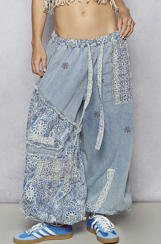 A person wearing loose, wide-leg light blue denim pants with patchwork floral lace panels and drawstring waist, paired with blue sneakers against a plain light gray wall.