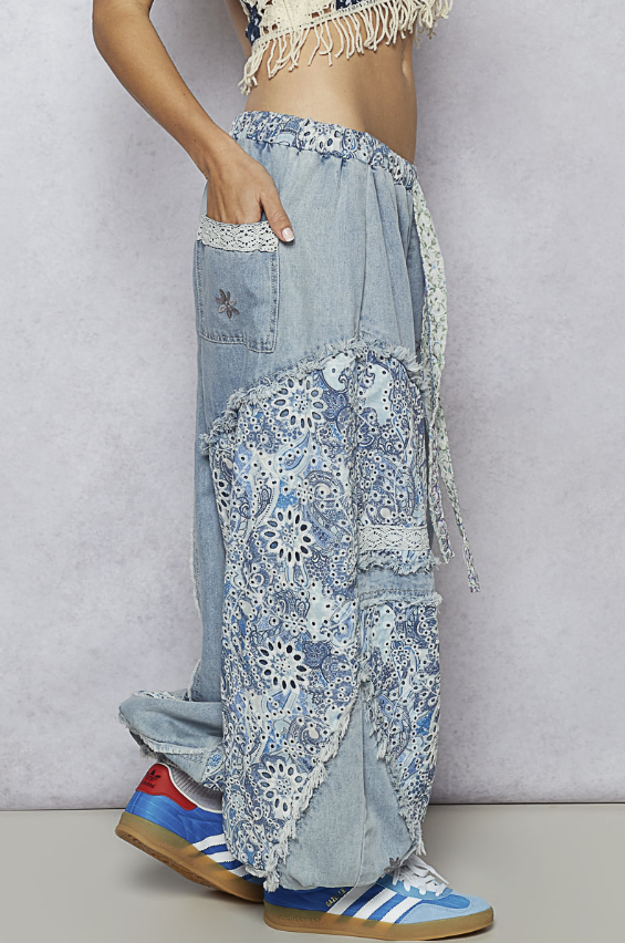 A person wearing light-wash blue denim jeans with extensive white floral crochet lace applique patches, side pockets, and a tie waist, paired with colorful sneakers against a textured gray wall.