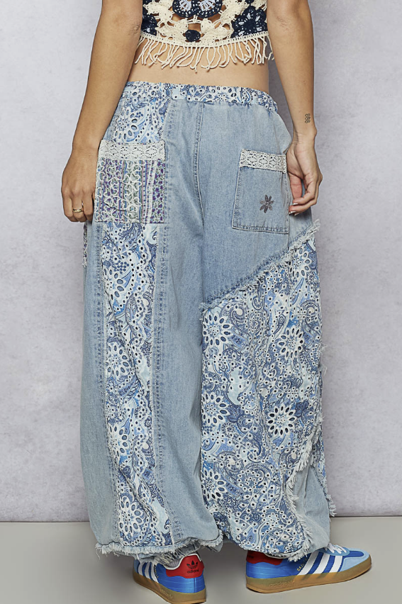 A person wearing loose, patchwork-style light blue denim pants with floral lace and embroidered panels, paired with blue and red sneakers against a plain light gray studio background.