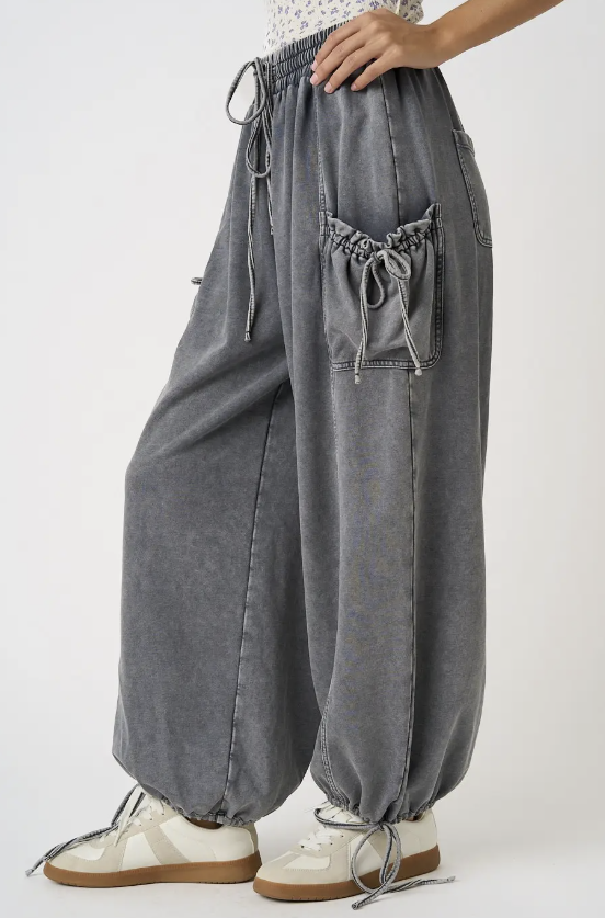 A person wearing loose, wide-leg gray drawstring pants with gathered cuffs and large front pockets, paired with white sneakers, photographed against a clean light gray studio background.