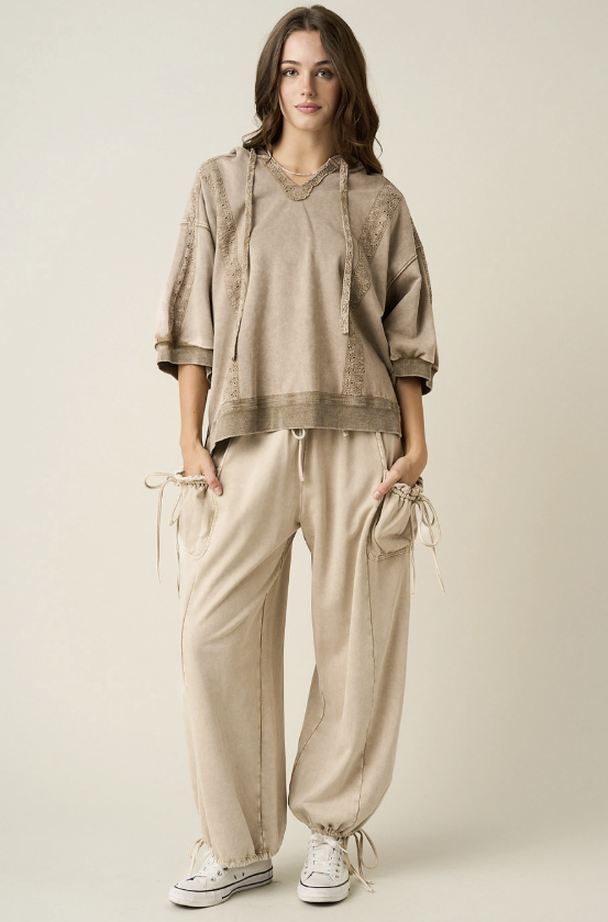 A woman modeling a matching beige lounge set consisting of a loose short-sleeve top with lace-up details and matching drawstring pants, paired with white sneakers, against a plain light beige studio background.