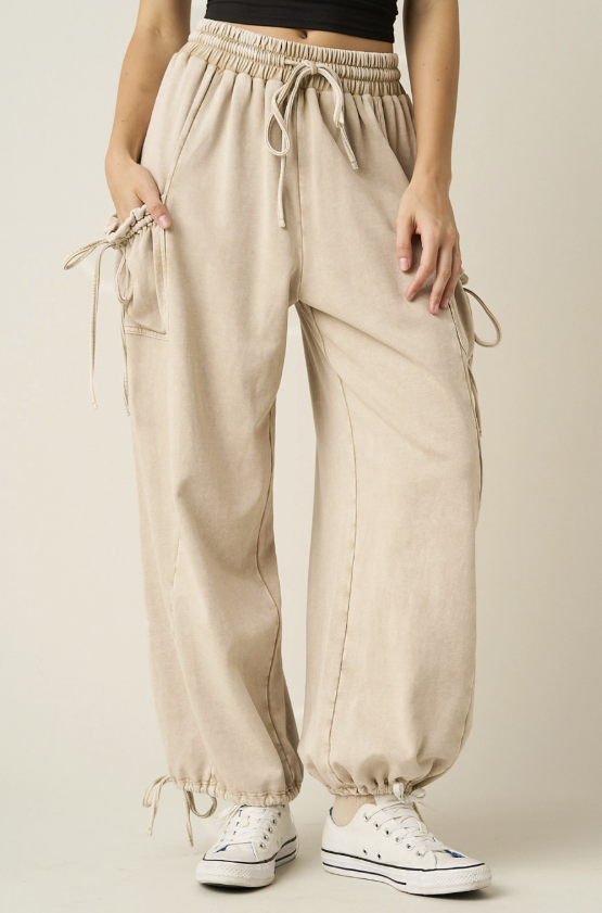 A person wearing loose beige drawstring jogger-style pants with side pockets and gathered cuffs, paired with white sneakers against a plain light beige studio background.