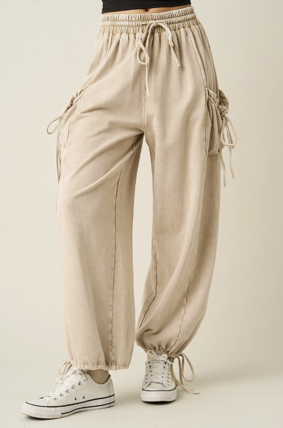 A person wearing loose beige drawstring pants with side cargo-style tie details and elastic ankle cuffs, paired with white low-top sneakers, against a plain light beige studio background.