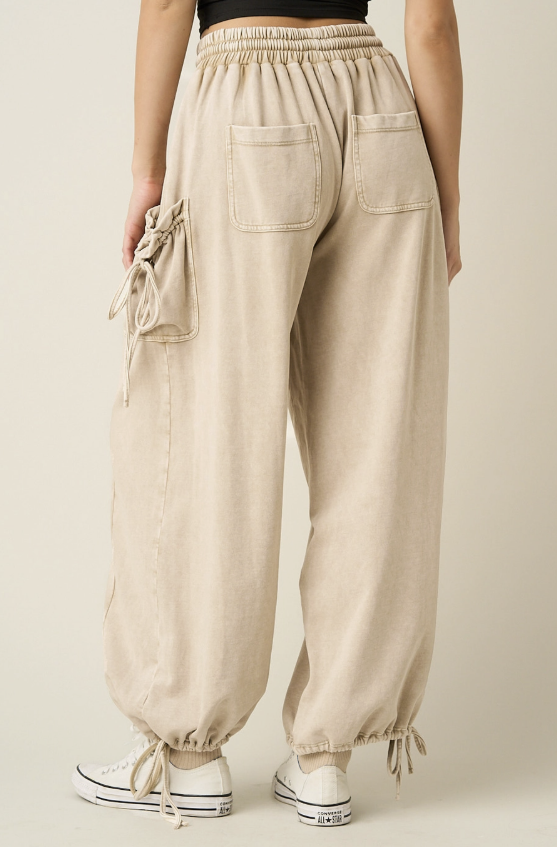 A person shown from the back wearing loose beige drawstring cargo-style pants with large back patch pockets and elastic cuffs, paired with white sneakers against a plain light beige studio background.