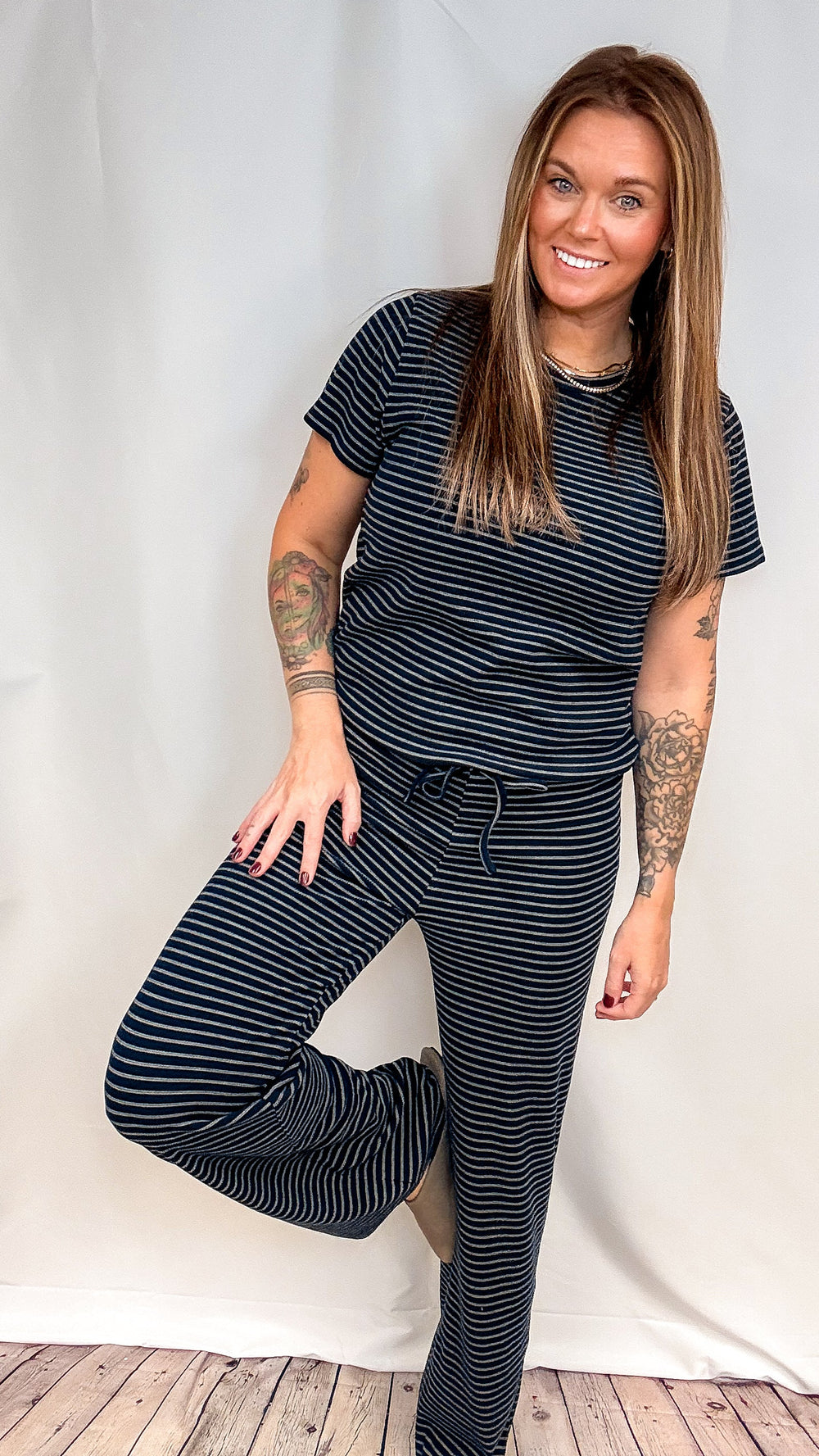 A woman modeling a matching two-piece loungewear set consisting of a short-sleeve t-shirt and drawstring pants, both in a navy blue and white stripe pattern, posed against a white fabric backdrop.