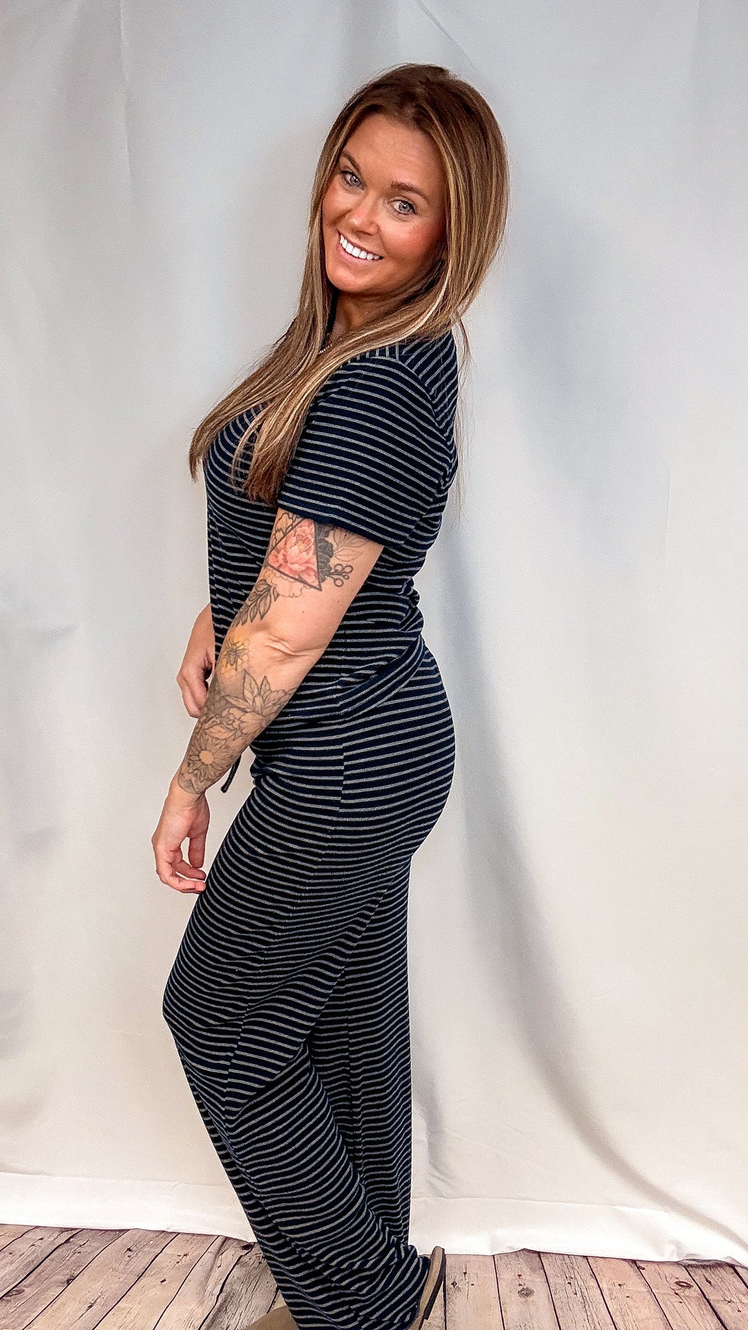 A woman modeling a navy blue and white horizontally striped short-sleeve jumpsuit, standing against a white fabric backdrop.