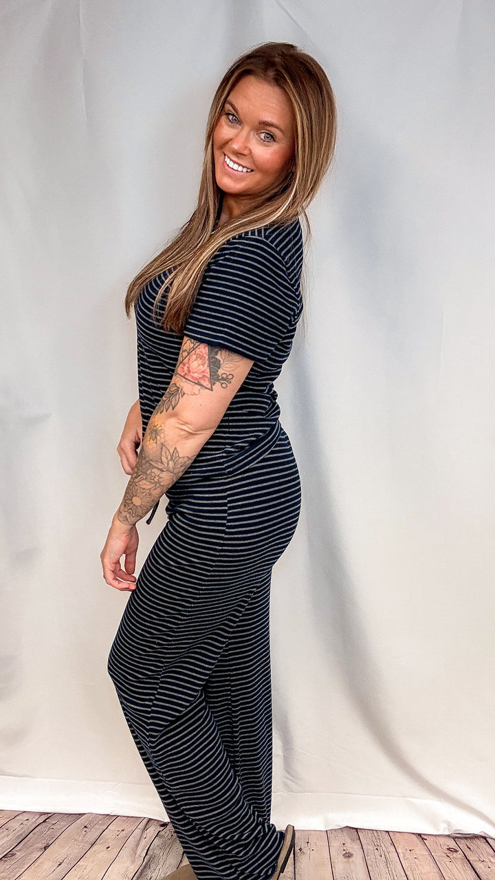 A woman modeling a navy blue and white horizontally striped short-sleeve jumpsuit, standing against a white fabric backdrop.