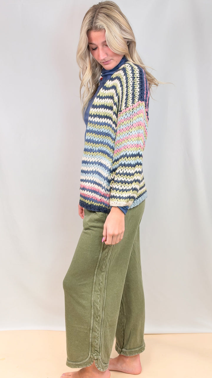 Woman shown from side wearing colorful stripe cardigan and green pants on plain background