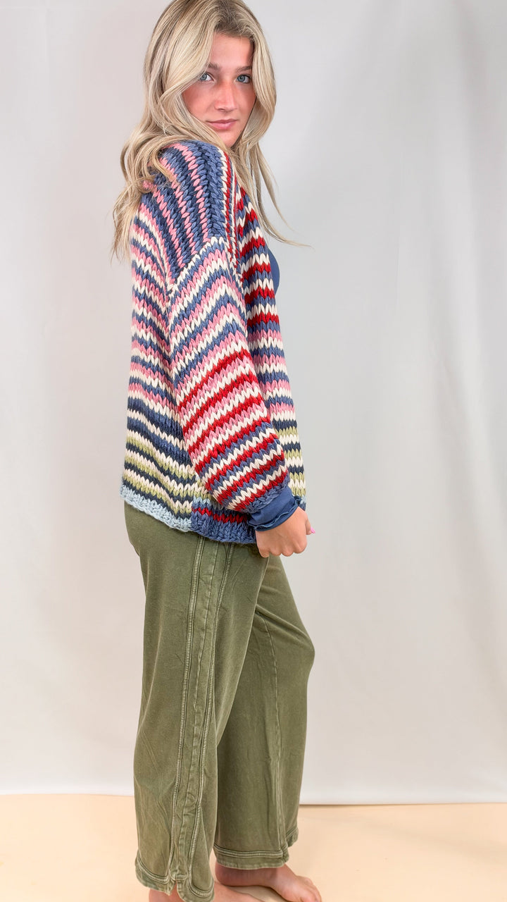 Woman shown from side view wearing colorful stripe cardigan and green pants on plain background