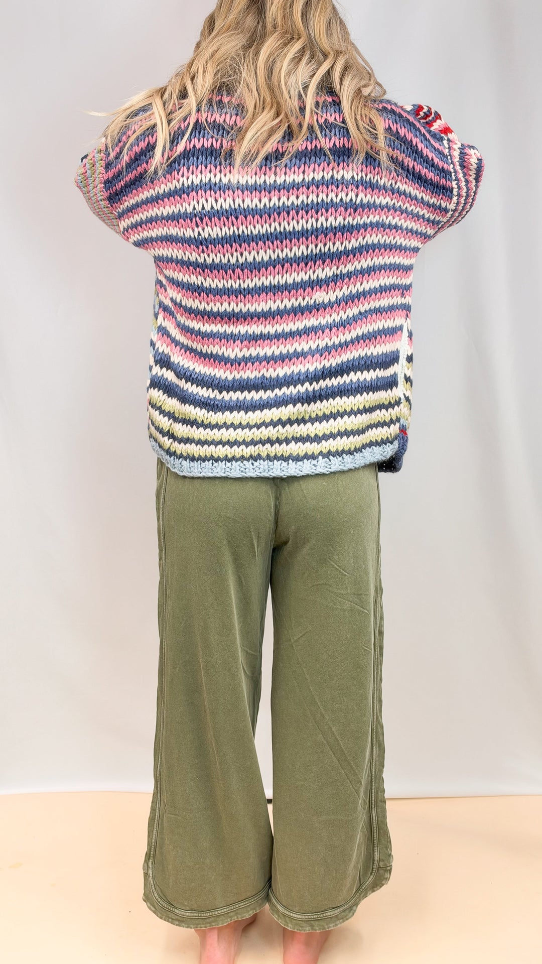 Woman shown from back view wearing colorful stripe sweater and green pants on plain background