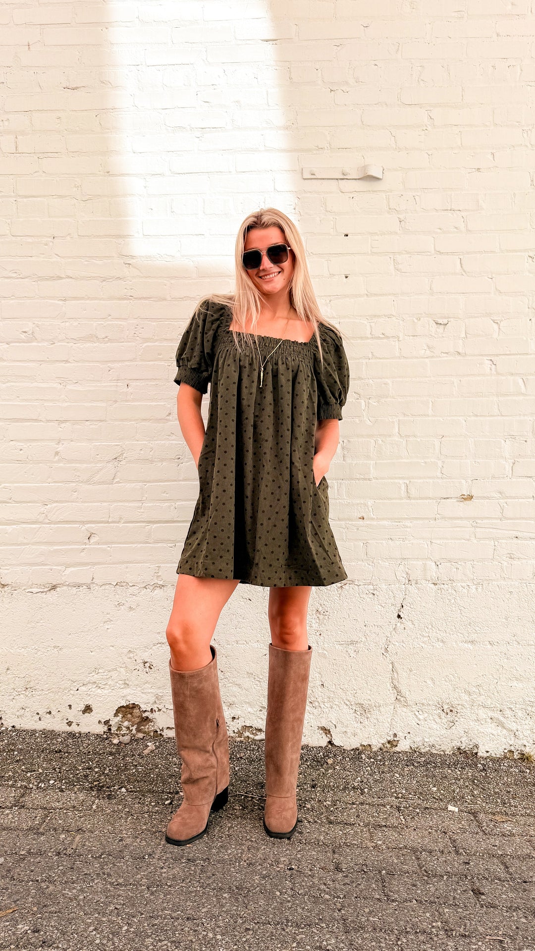 Woman wearing olive mini dress with brown polka dots and tall beige boots in front of white brick wall