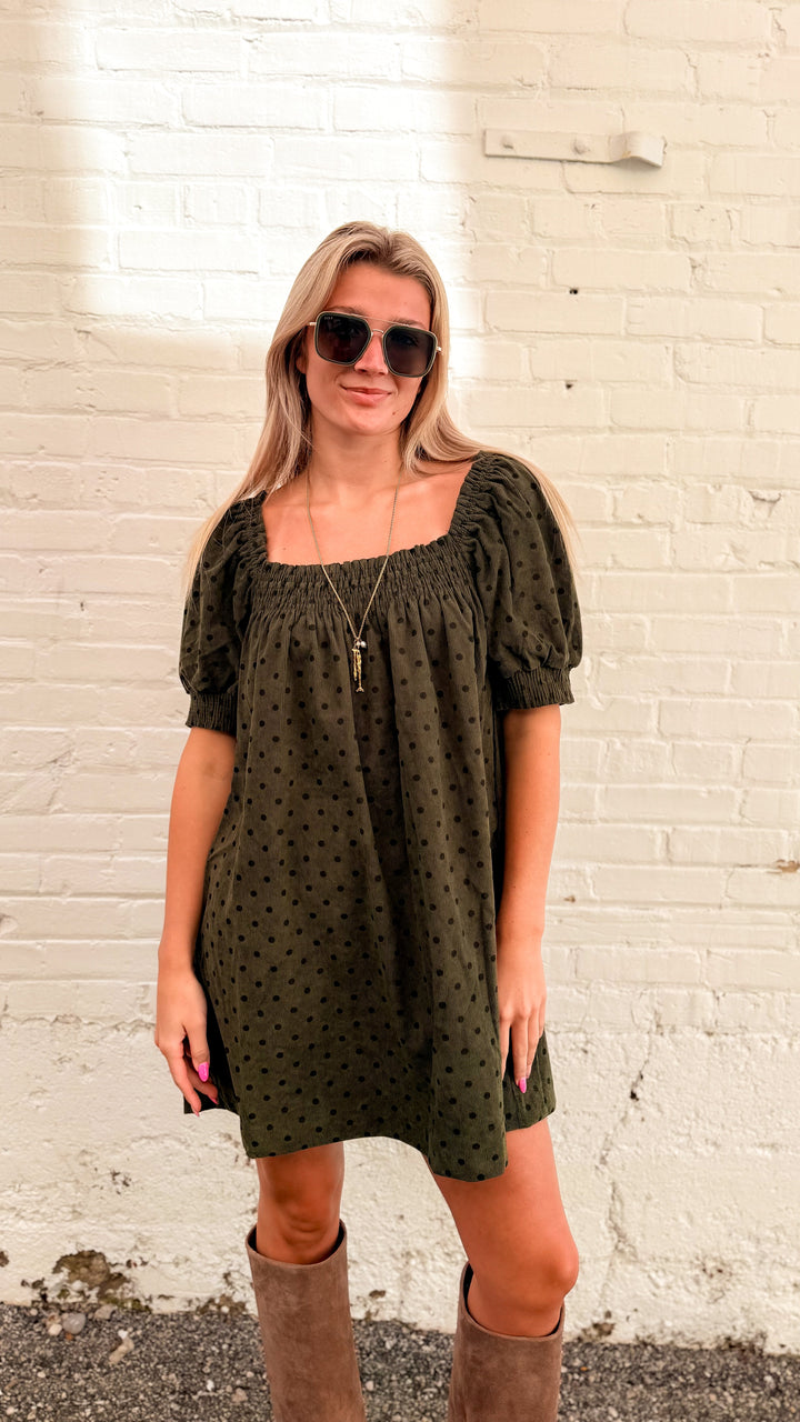 Woman wearing olive green mini dress with brown polka dots and tall beige boots in front of white brick wall