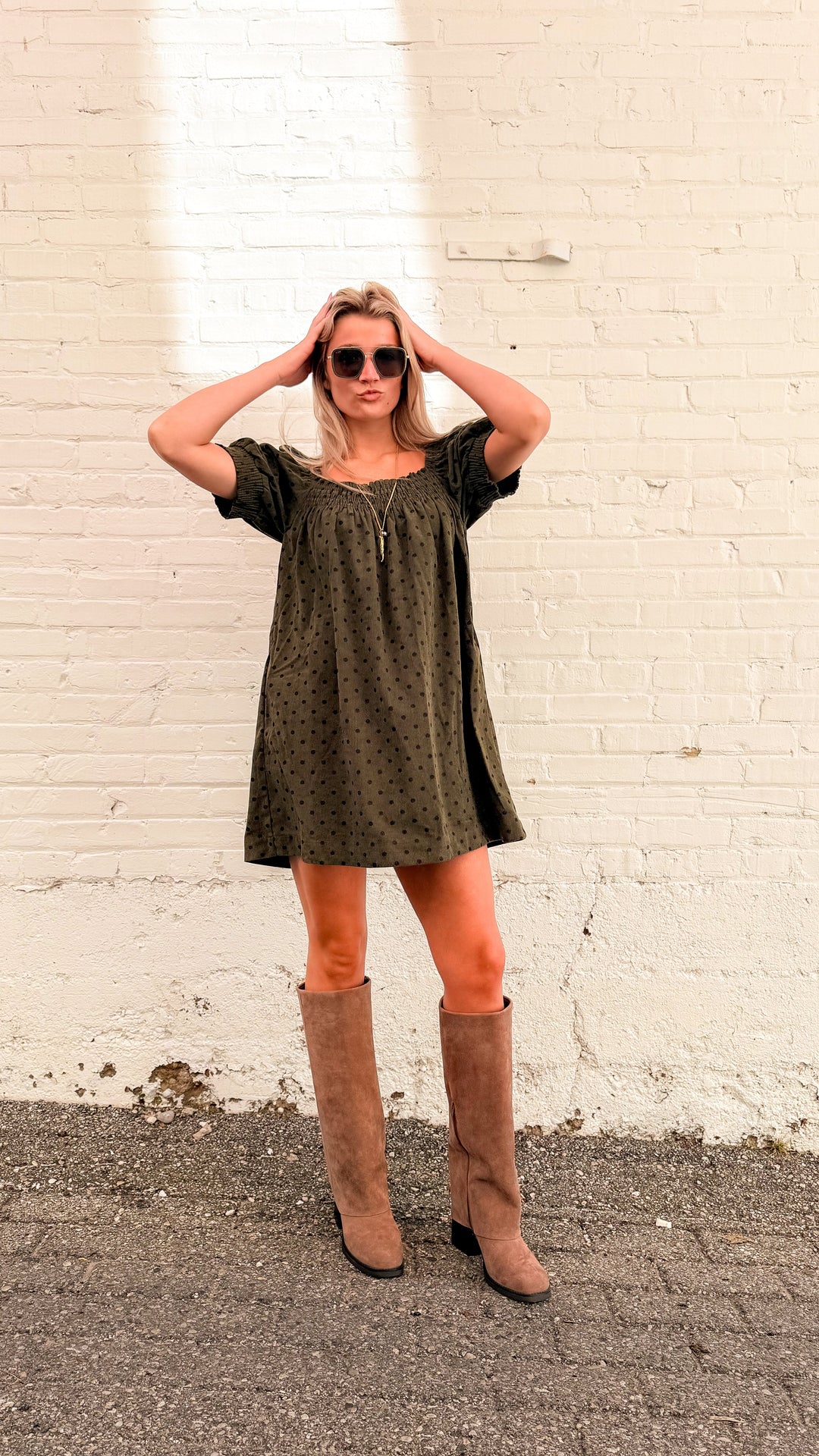 Woman wearing olive green dress with brown polka dots and tall beige boots in front of white brick wall