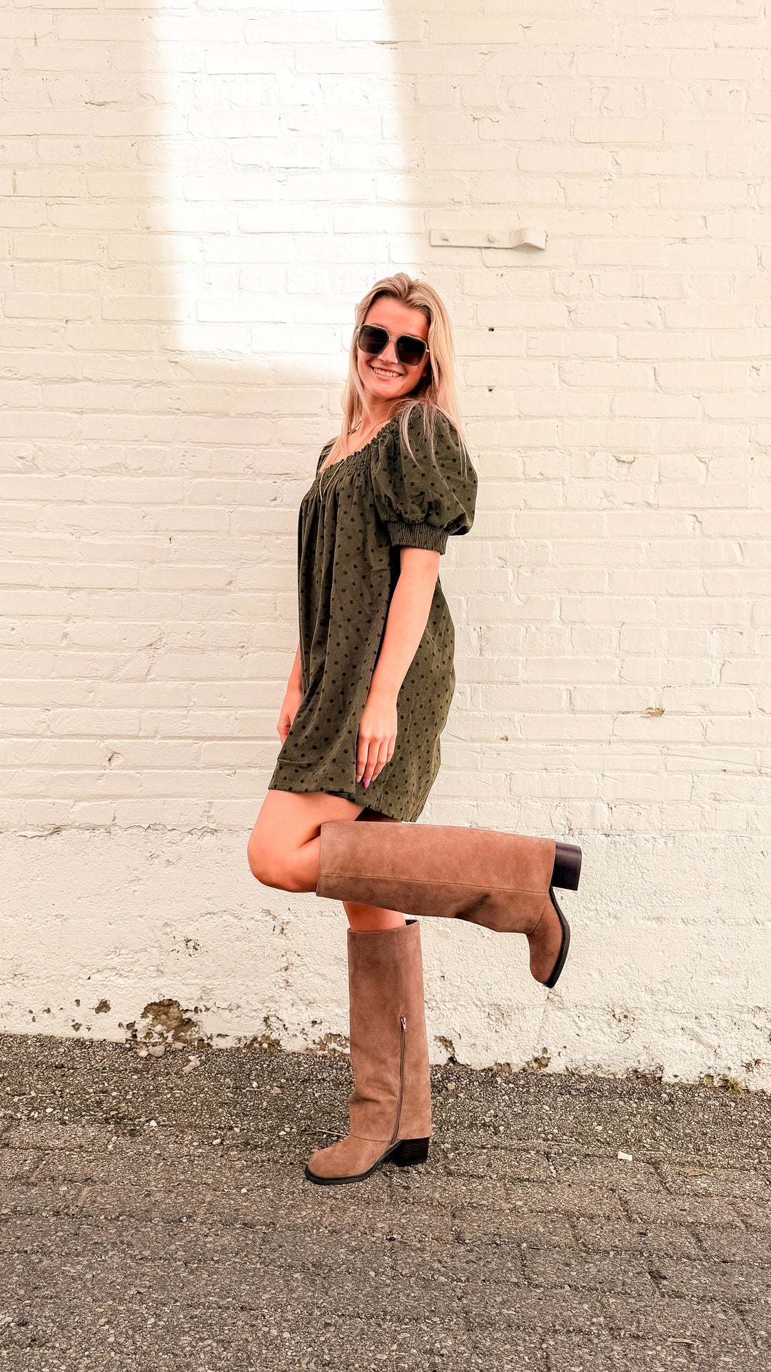 Woman wearing olive green mini dress with brown polka dots and tall beige boots in front of white brick wall 