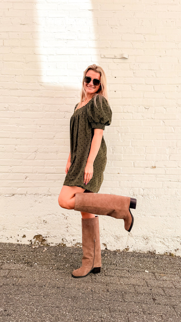 Woman wearing olive green mini dress with brown polka dots and tall beige boots in front of white brick wall 