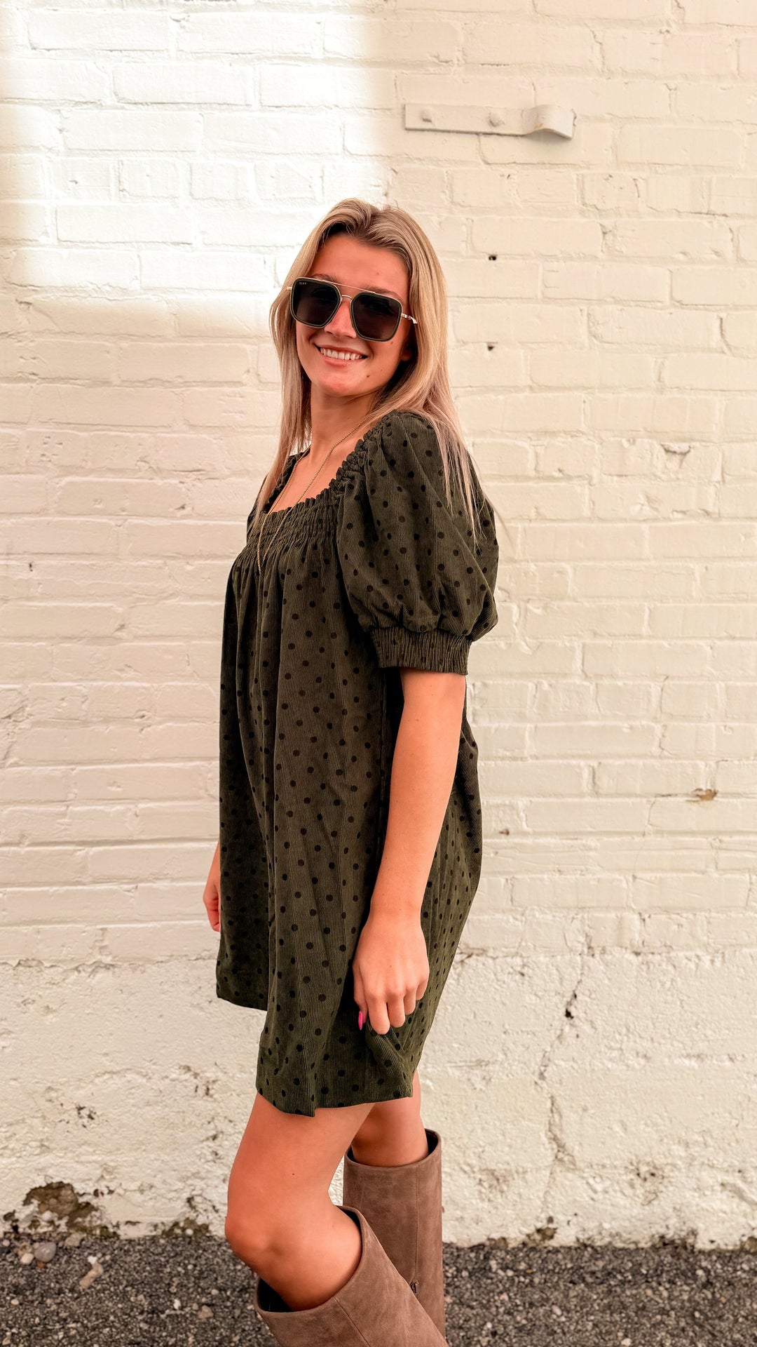 Woman wearing olive green mini dress with brown polka dots and tall beige boots in front of white brick wall