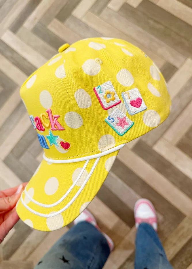 Yellow polka dot cap with colorful patches held by a person on a wooden floor.