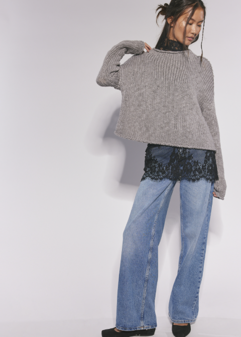 A model wearing a gray ribbed knit cropped sweater, blue wide-leg jeans, and black lace hem detail, standing against a plain light gray studio background.