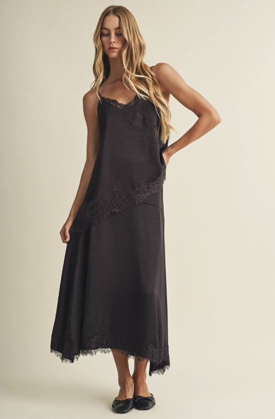 A woman modeling a brown sleeveless maxi slip dress with lace trim and a draped, gathered silhouette, photographed against a plain warm beige studio background.