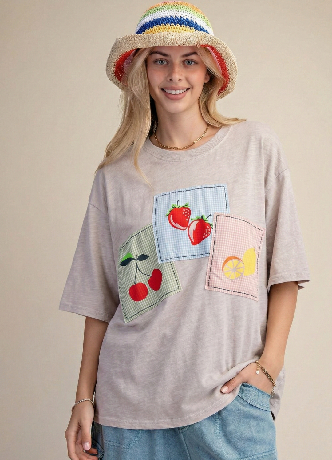 Woman wearing a gray t-shirt with colorful fruit patches and a rainbow hat on a beige background