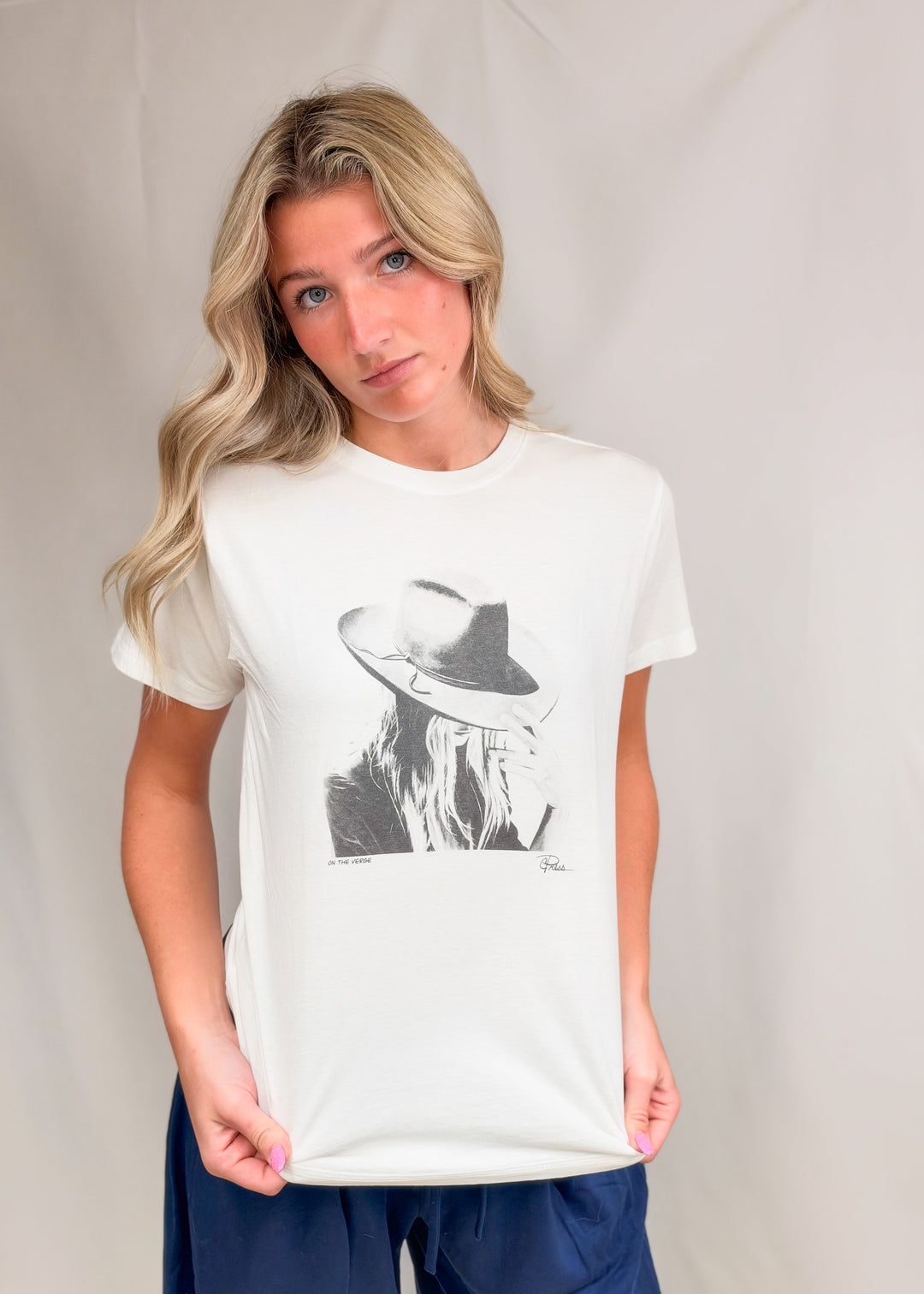 Woman wearing graphic tee with persons face and hat with blue pants on plain background