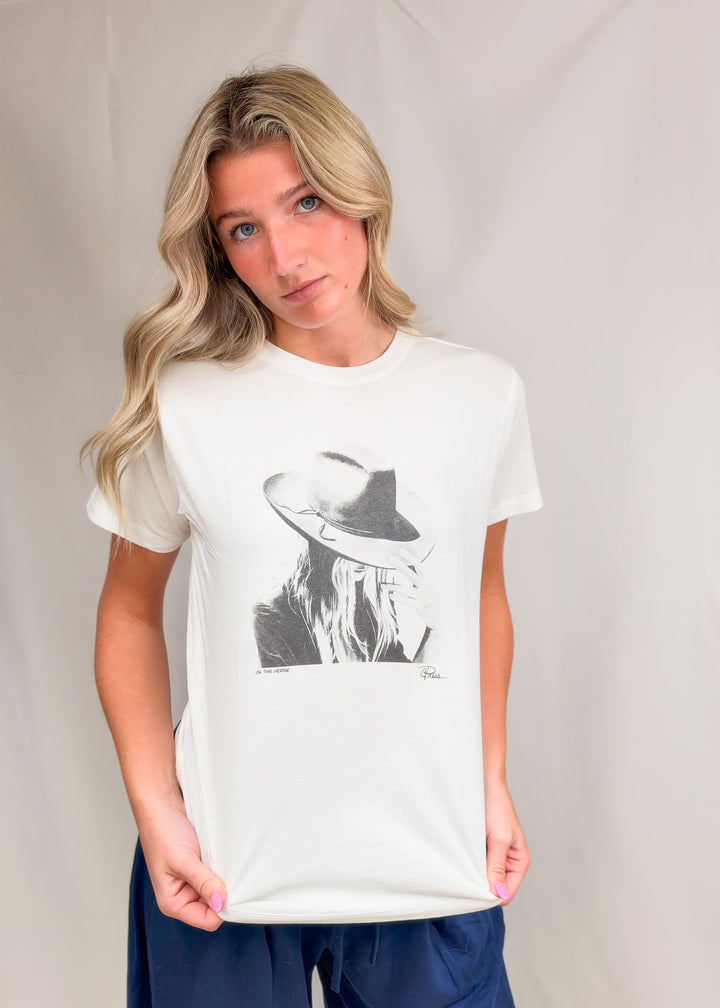 Woman wearing graphic tee with persons face and hat with blue pants on plain background