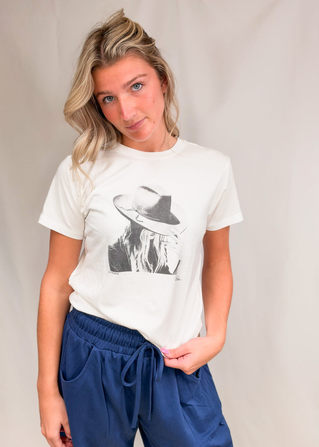 Woman wearing graphic tee with persons face wearing a hat with blue pants on plain background