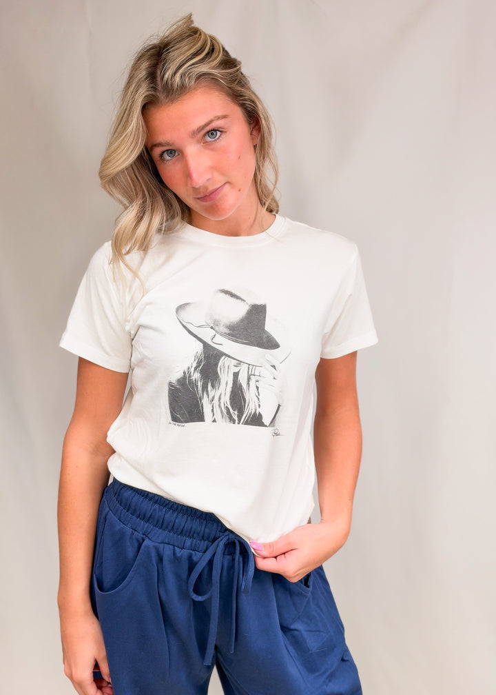 Woman wearing graphic tee with persons face wearing a hat with blue pants on plain background