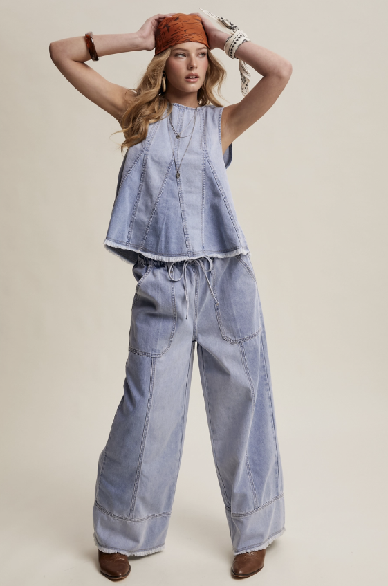 A woman modeling a light blue denim two-piece set consisting of a sleeveless cropped top and wide-leg pants, styled with a rust-colored bandana, bracelets, and brown boots against a solid beige background.